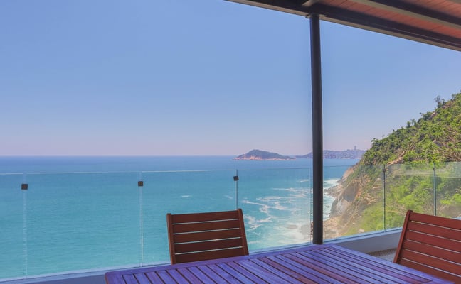 Covered terrace with wooden table and chairs overlooking turquoise sea, rocky coastline, and distant island