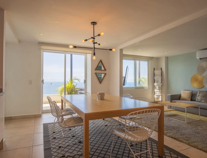 Open-plan living and dining area with ocean view, wooden dining table, woven chairs, and modern pendant lighting